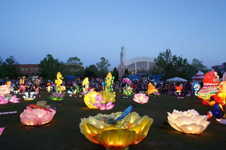 The image depicts the International Night Market in Suwanee's "Glow in the Park" lantern parade, featuring a variety of illuminated, colorful lanterns arranged on a grassy area. The lanterns are shaped like vibrant flowers, animals, and abstract figures, creating a lively and festive atmosphere.